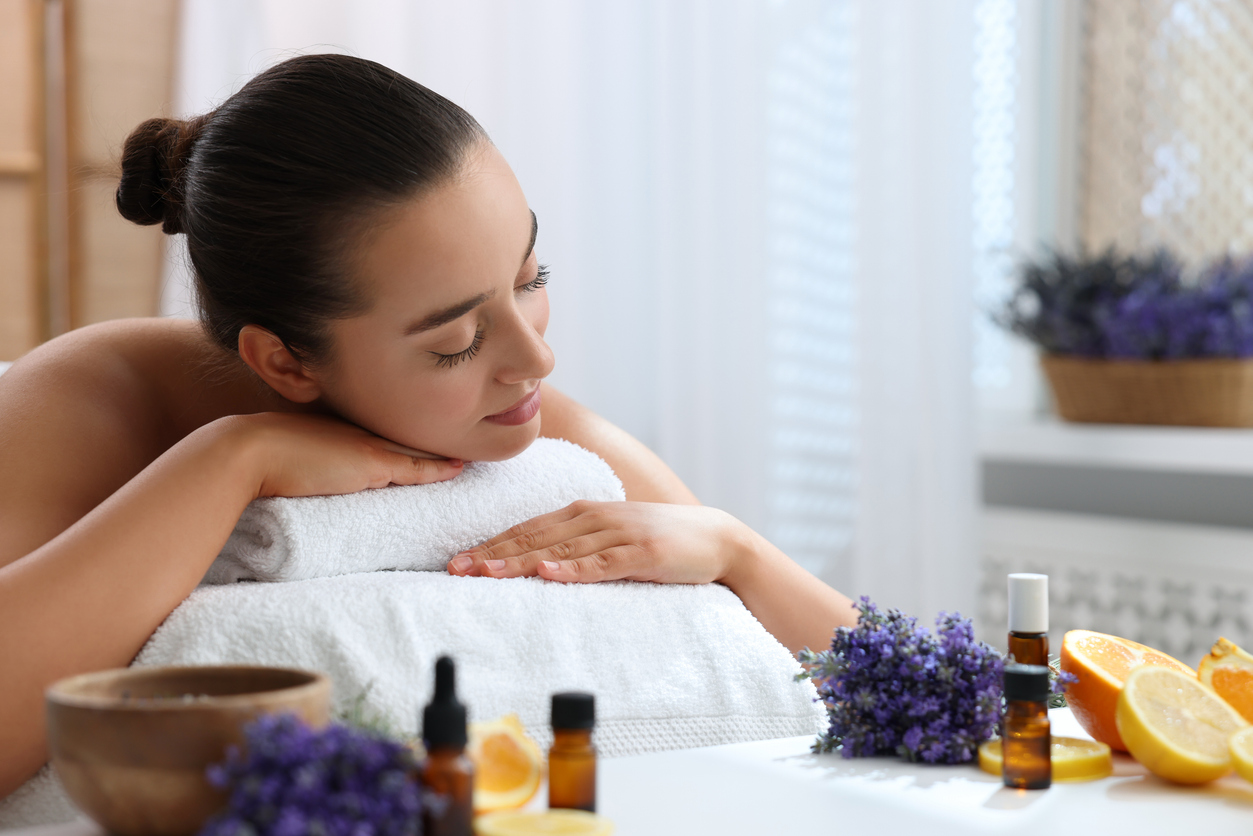 Beautiful young woman relaxing on massage couch and bottles of essential oil with ingredients on table in spa salon Beautiful young woman relaxing on massage couch and bottles of essential oil with ingredients on table in spa salon