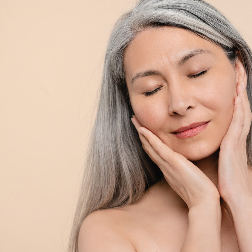Closeup cropped mature middle-aged woman with grey hair and eyes closed naked shirtless isolated in beige background. Beautification, rejuvenation, pampering concept. Skin and hair care