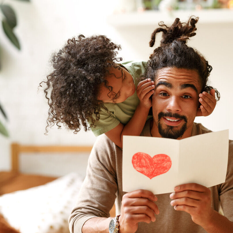 Smiling afro american boy son covering eyes of father with hands and congratulating him on holiday Surprise for daddy. Cute smiling afro american kid son giving father postcard with red drawn heart and covering his eyes with hands, happy child congratulating dad during family holiday at home