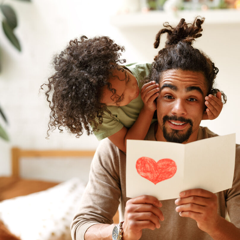 Surprise for daddy. Cute smiling afro american kid son giving father postcard with red drawn heart and covering his eyes with hands, happy child congratulating dad during family holiday at home