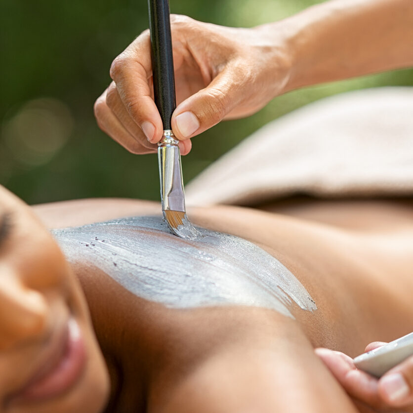 Hands of masseuse applying mud mask on african woman back. Young african american girl relaxing with eyes closed while beautician applying exfoliation clay on shoulder. Woman at resort doing clay mask exfoliation.