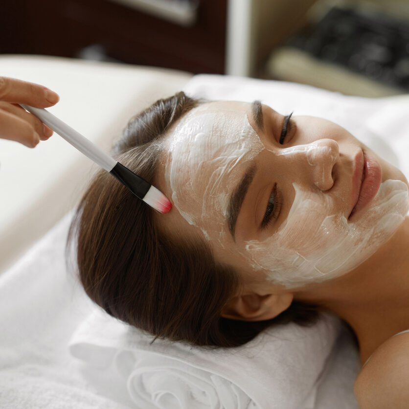 Facial Beauty Treatment. Closeup Of Beautiful Smiling Woman Getting Mask At Spa Salon. Cosmetologist Applying Cosmetic Mask With Brush On Female Face Indoors. Skin Care Concept. High Resolution Image